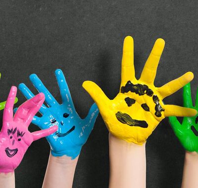 Bunte Kinderhände mit aufgemalten Gesichtern vor Tafel mit gezeichneten Wolken hochgestreckt